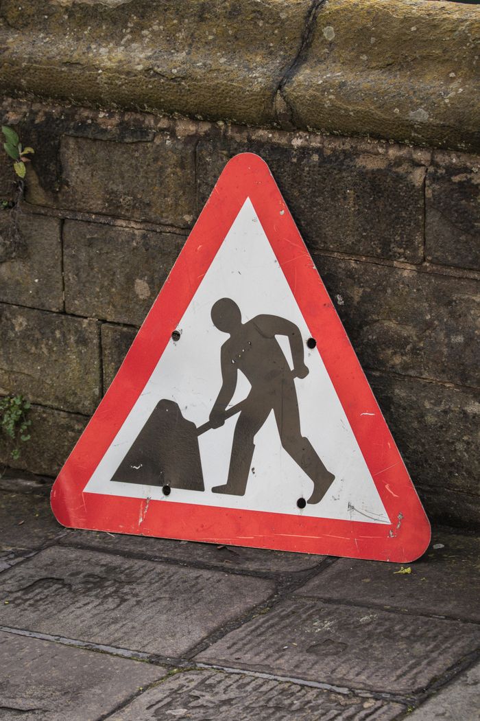 Triangular roadwork sign leaning against a stone wall, symbolizing construction ahead.